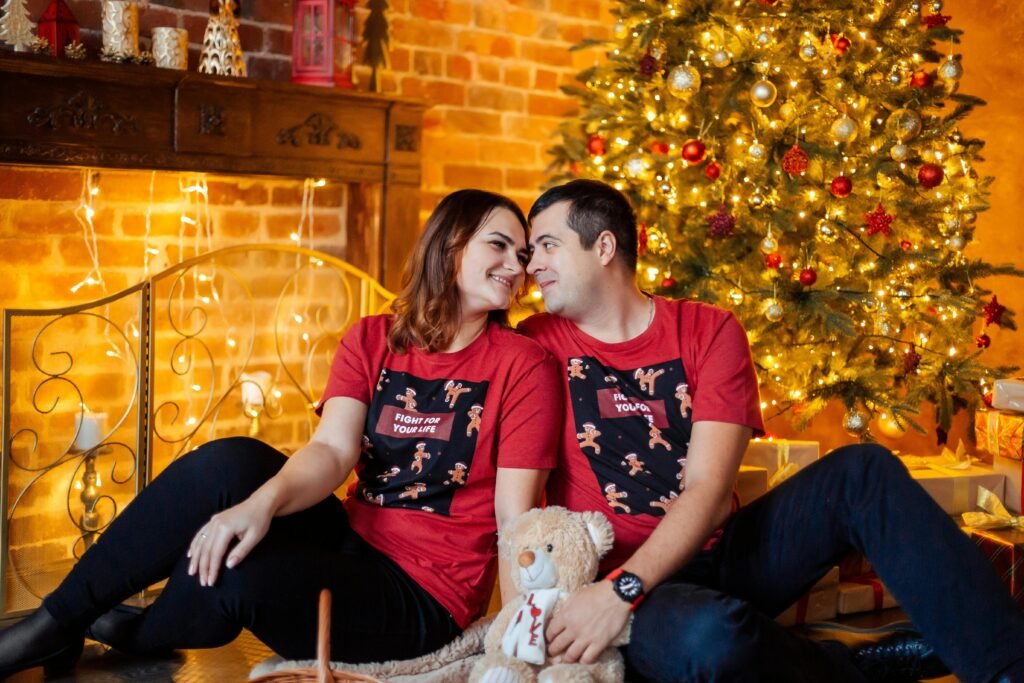 A smiling couple in red shirts enjoys Christmas by a decorated tree with lights and gifts.