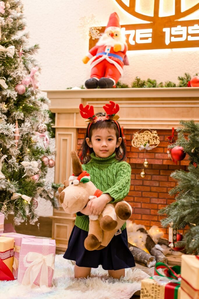 Child in festive attire holding a reindeer stuffed toy by the Christmas tree.