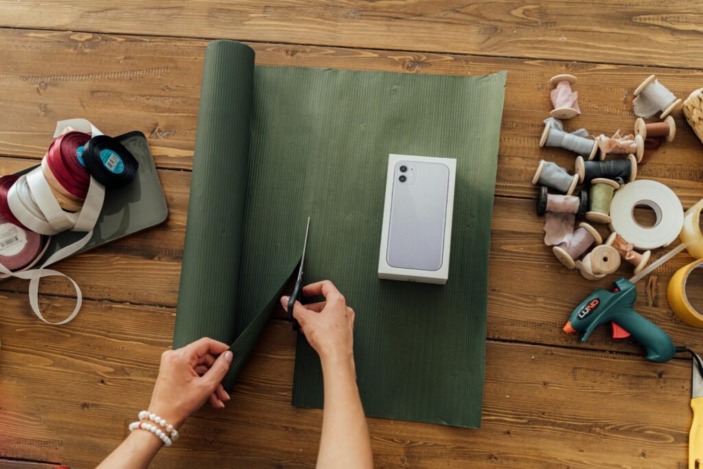 Hands wrapping a smartphone box with green paper on a wooden table, surrounded by ribbons and tools.