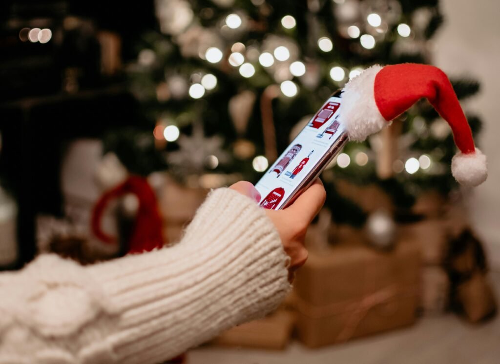 Close-up of a hand holding a smartphone with a Santa hat near a Christmas tree indoors.