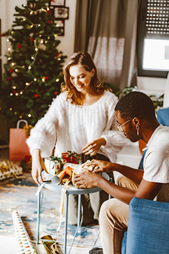 Two friends joyfully wrap gifts near a Christmas tree, capturing a warm festive atmosphere indoors.