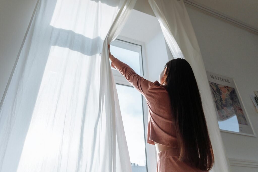 Woman opens white curtains in bright room, revealing a sunny day outside.